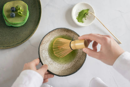 Person using a bamboo whisk to mix green tea in a ceramic bowl on a white surface.