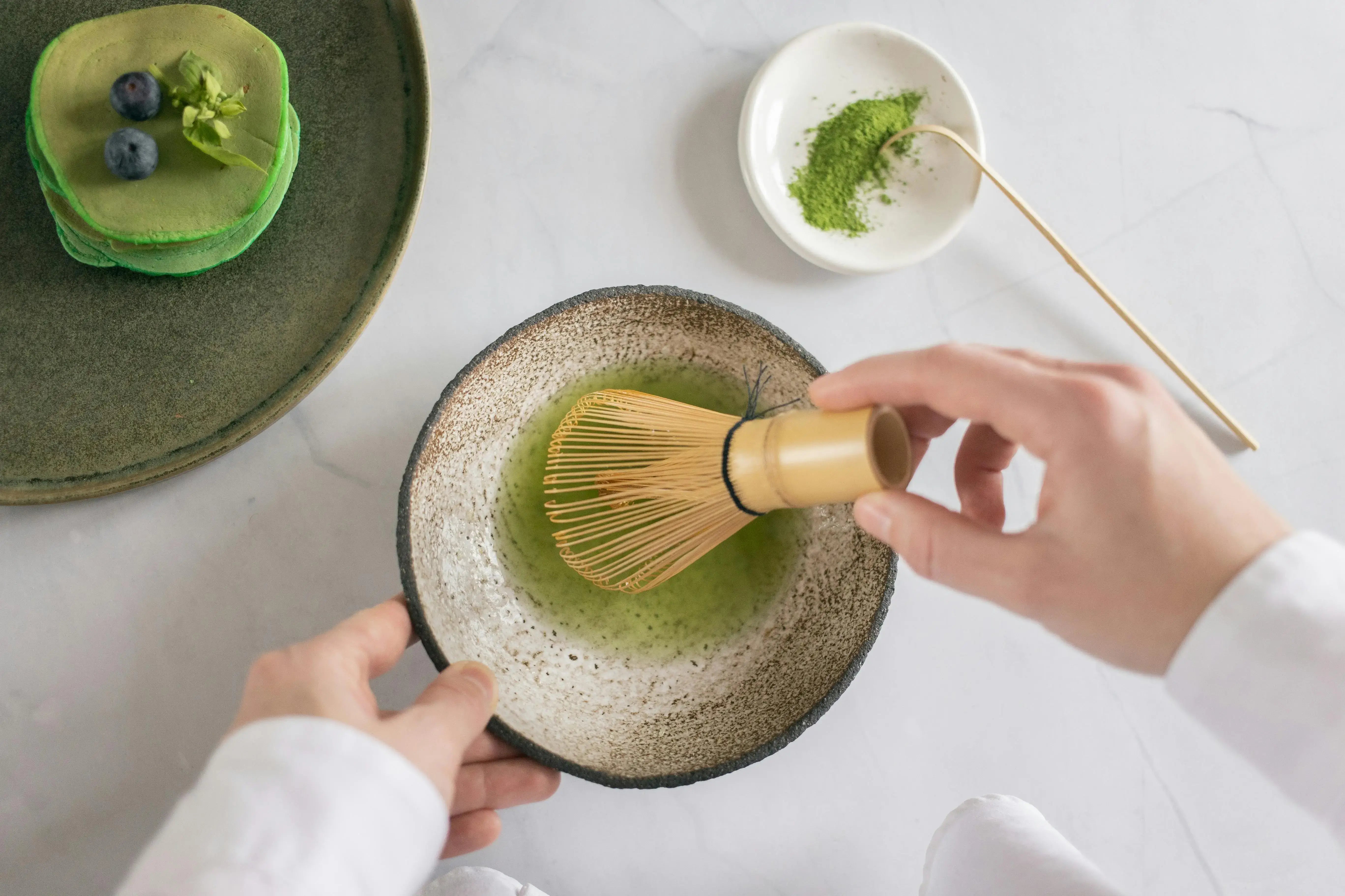 Person using a bamboo whisk to mix green tea in a ceramic bowl on a white surface.