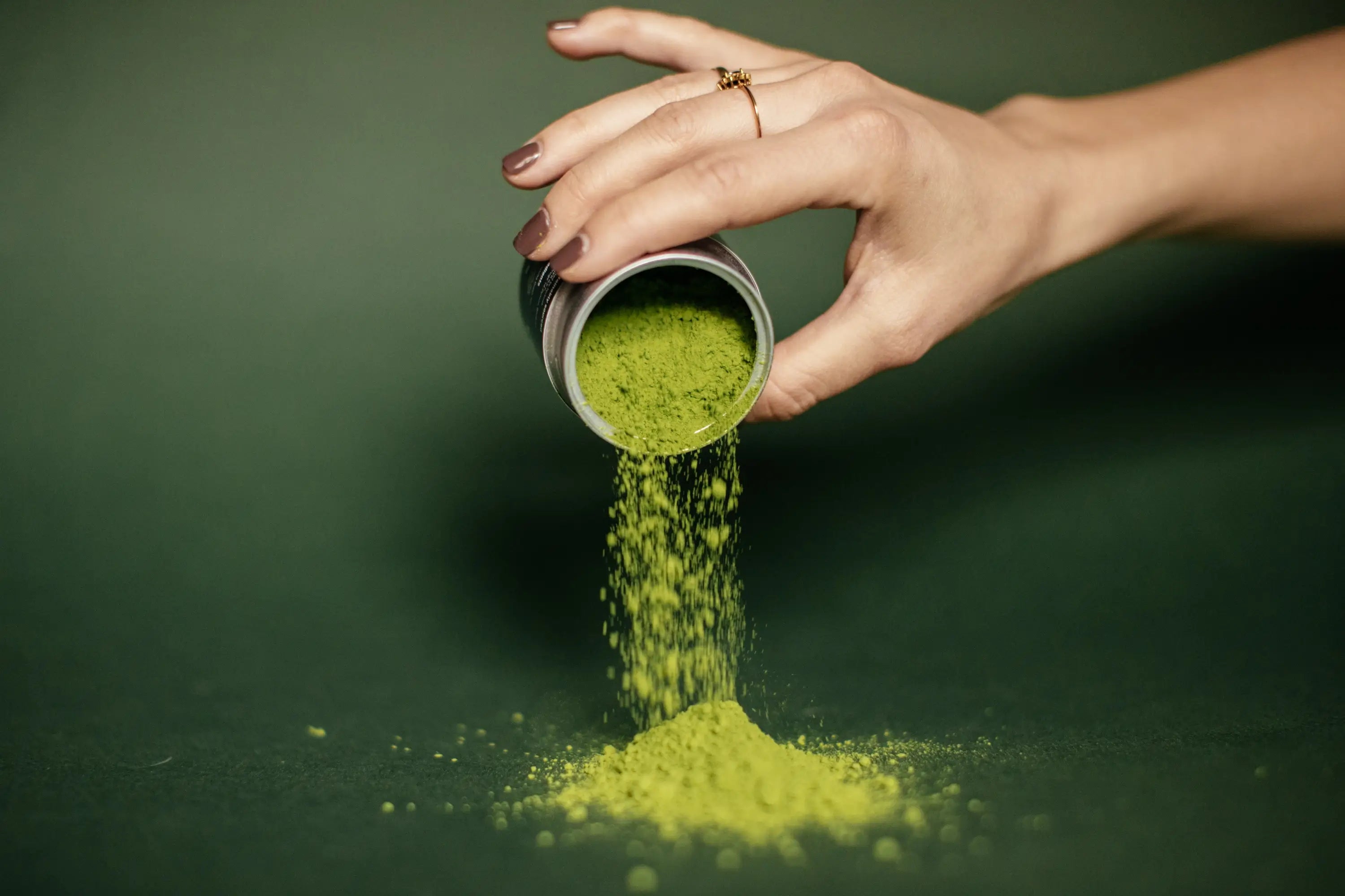 Hand pouring green powder from a small container onto a dark green background